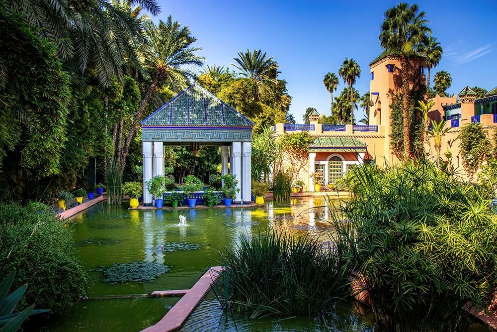 Maison de Jardin Majorelle. Green pond in the foreground, gazebo in the center, and a beautiful terracota and blue building on the right side of the photo.