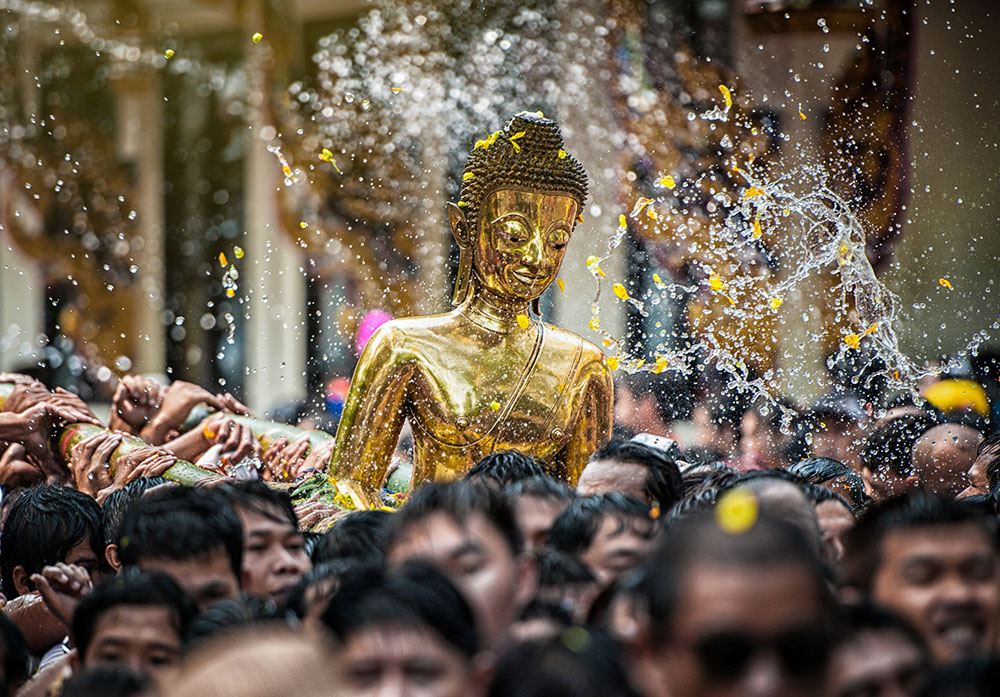 Crowd of people marching with large Buddha statue and water being thrown in the background.